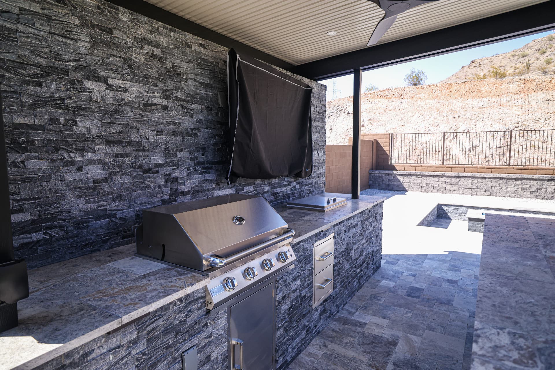 Covered outdoor kitchen with stacked stone bar, grill, and TV